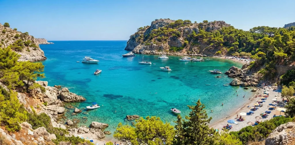 A scenic panoramic view of Anthony Quinn Bay in Rhodes, Greece, featuring crystal-clear turquoise waters, anchored boats, and sunbathers relaxing on the pebbled beach surrounded by rocky cliffs.